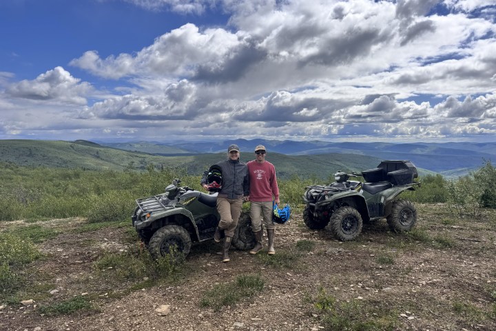 Two people with ATVs on scenic mountain trail under cloudy sky.