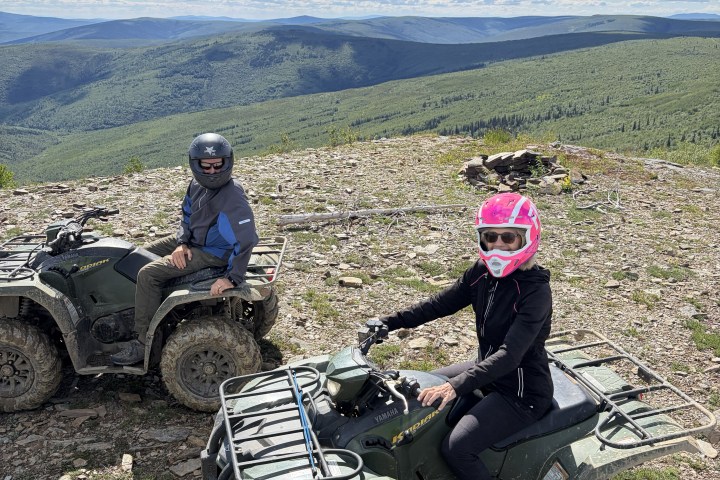 Two people on ATVs with helmets atop a hill overlooking a vast landscape and cloudy sky.