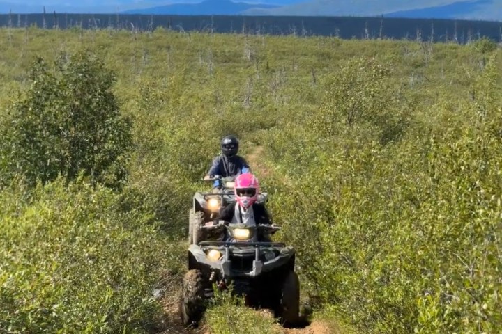 Two people riding ATVs on a narrow trail through dense green vegetation.