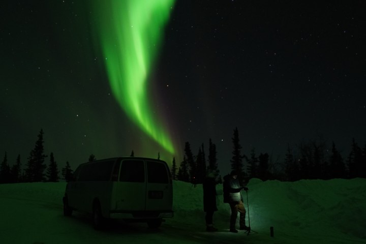 Green aurora borealis over snowy landscape with van and people.