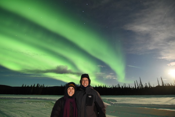 Two people smiling under vibrant green northern lights in a snowy landscape.